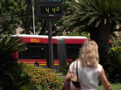 Eine Frau läuft neben einem Bus vorbei auf dem das Wort Sevilla steht. Über ihrem Kopf zeigt ein Thermometer die Temperatur von 44 Grad Celsius an.