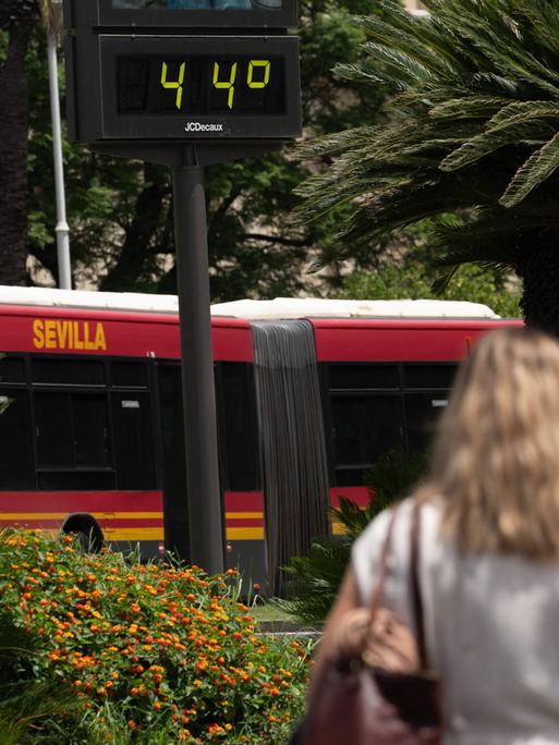 Eine Frau läuft neben einem Bus vorbei auf dem das Wort Sevilla steht. Über ihrem Kopf zeigt ein Thermometer die Temperatur von 44 Grad Celsius an.