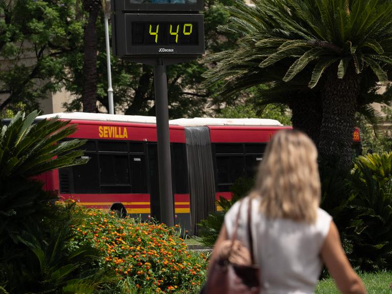 Eine Frau läuft neben einem Bus vorbei auf dem das Wort Sevilla steht. Über ihrem Kopf zeigt ein Thermometer die Temperatur von 44 Grad Celsius an.