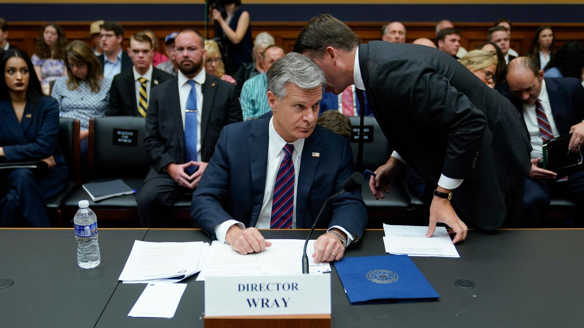 Der grauhaarige Wray sitzt hinter einem Mikrofon an einem Tisch. Ein Mitarbeiter steht neben ihm und sagt ihm etwas in Ohr. Dahinter Sitzreiehn mit Zuhörern. Der grauhaarige Wray sitzt hinter einem Mikrofon an einem Tisch. Ein Mitarbeiter steht neben ihm und sagt ihm etwas in Ohr. Dahinter Sitzreiehn mit Zuhörern.
