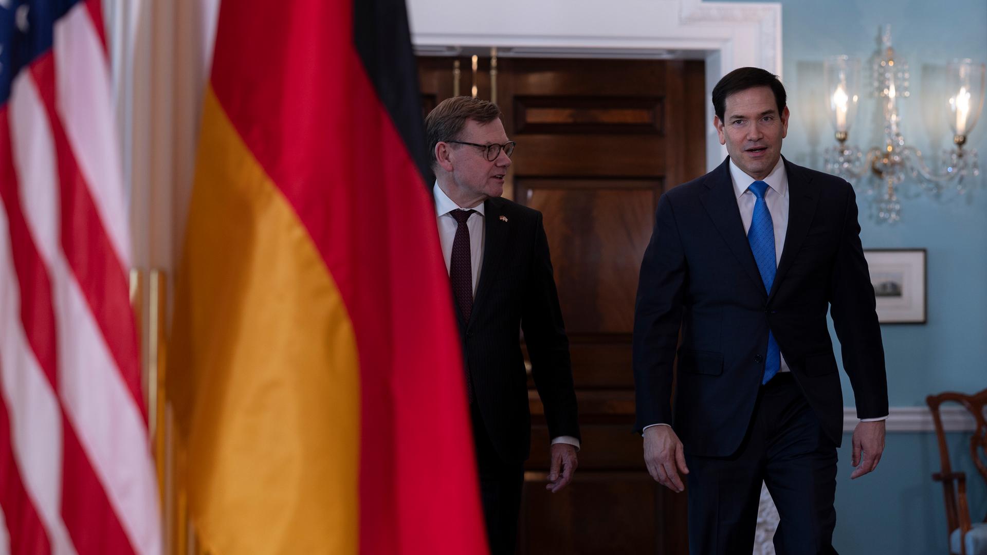 Bundesaußenminister Johann Wadephul (CDU) geht neben seinem Amtskollegen, US-Außenminister Marco Rubio, durch die Räume des US-Außenministeriums. Im Vordergrund sind die deutsche und die amerikanische Flagge im Bild.