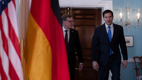 Bundesaußenminister Johann Wadephul (CDU) geht neben seinem Amtskollegen, US-Außenminister Marco Rubio, durch die Räume des US-Außenministeriums. Im Vordergrund sind die deutsche und die amerikanische Flagge im Bild.