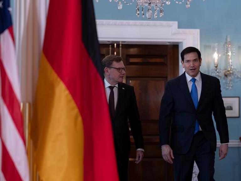 Bundesaußenminister Johann Wadephul (CDU) geht neben seinem Amtskollegen, US-Außenminister Marco Rubio, durch die Räume des US-Außenministeriums. Im Vordergrund sind die deutsche und die amerikanische Flagge im Bild.
