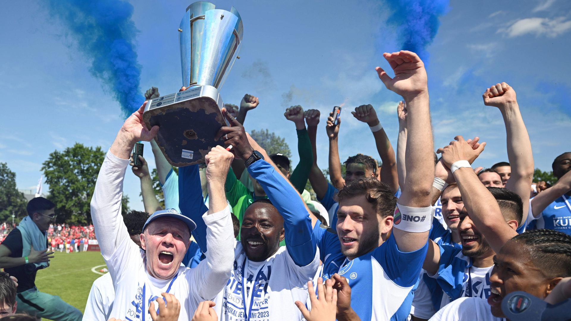 Mehrere Fußball-Spieler jubeln nach einem Sieg gemeinsam mit Fans und ihrem Trainer auf dem Spielfeld und halten einen Pokal in die Höhe.