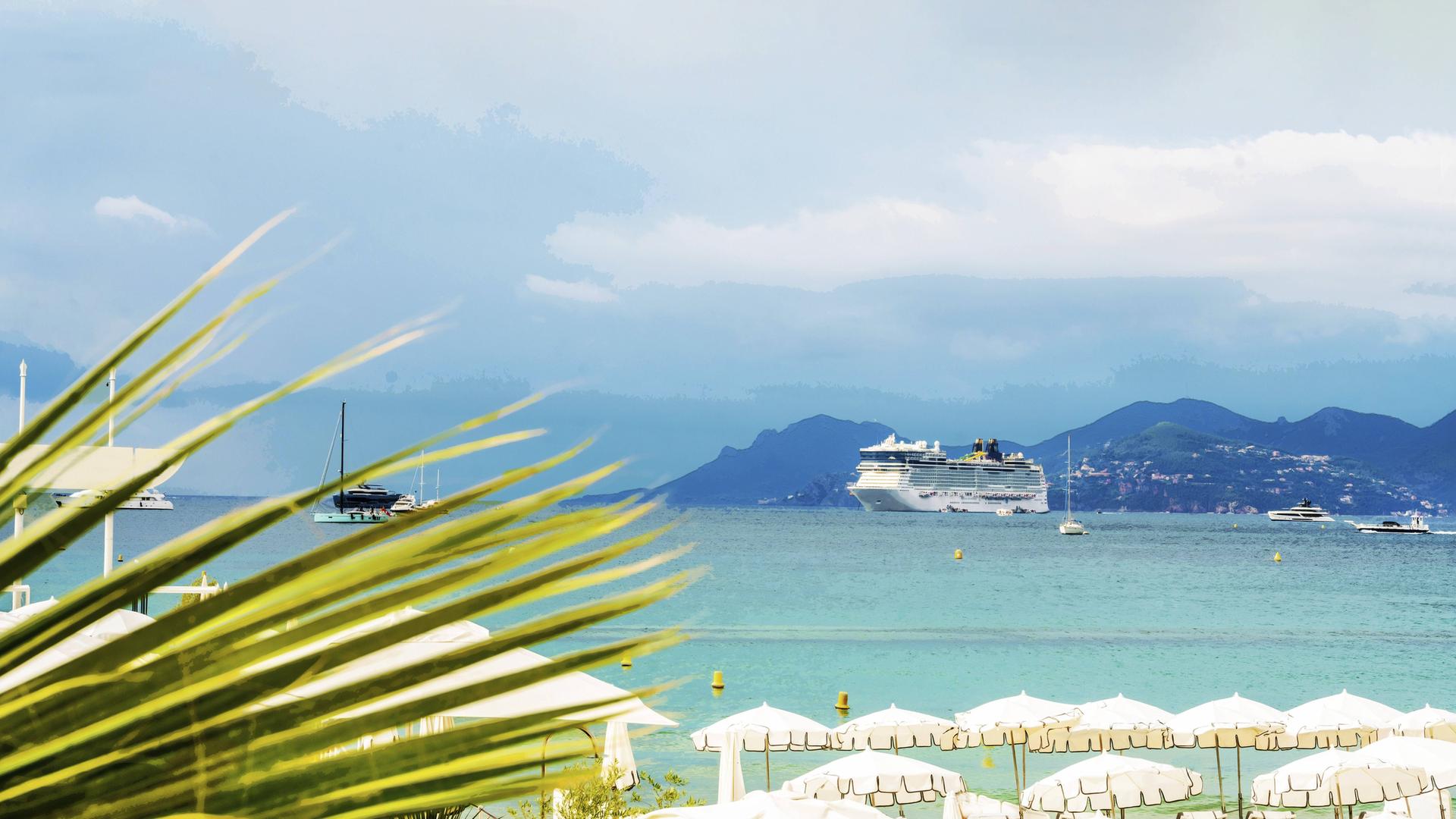 Ein Kreuzfahrtschiff in der Bucht von Cannes. Im Vordergrund sind Sonnenschirme an einem Strand zu sehen. Im Hintergrund eine Stadt in den Bergen. Ein Kreuzfahrtschiff in der Bucht von Cannes. Im Vordergrund sind Sonnenschirme an einem Strand zu sehen. Im Hintergrund eine Stadt in den Bergen.