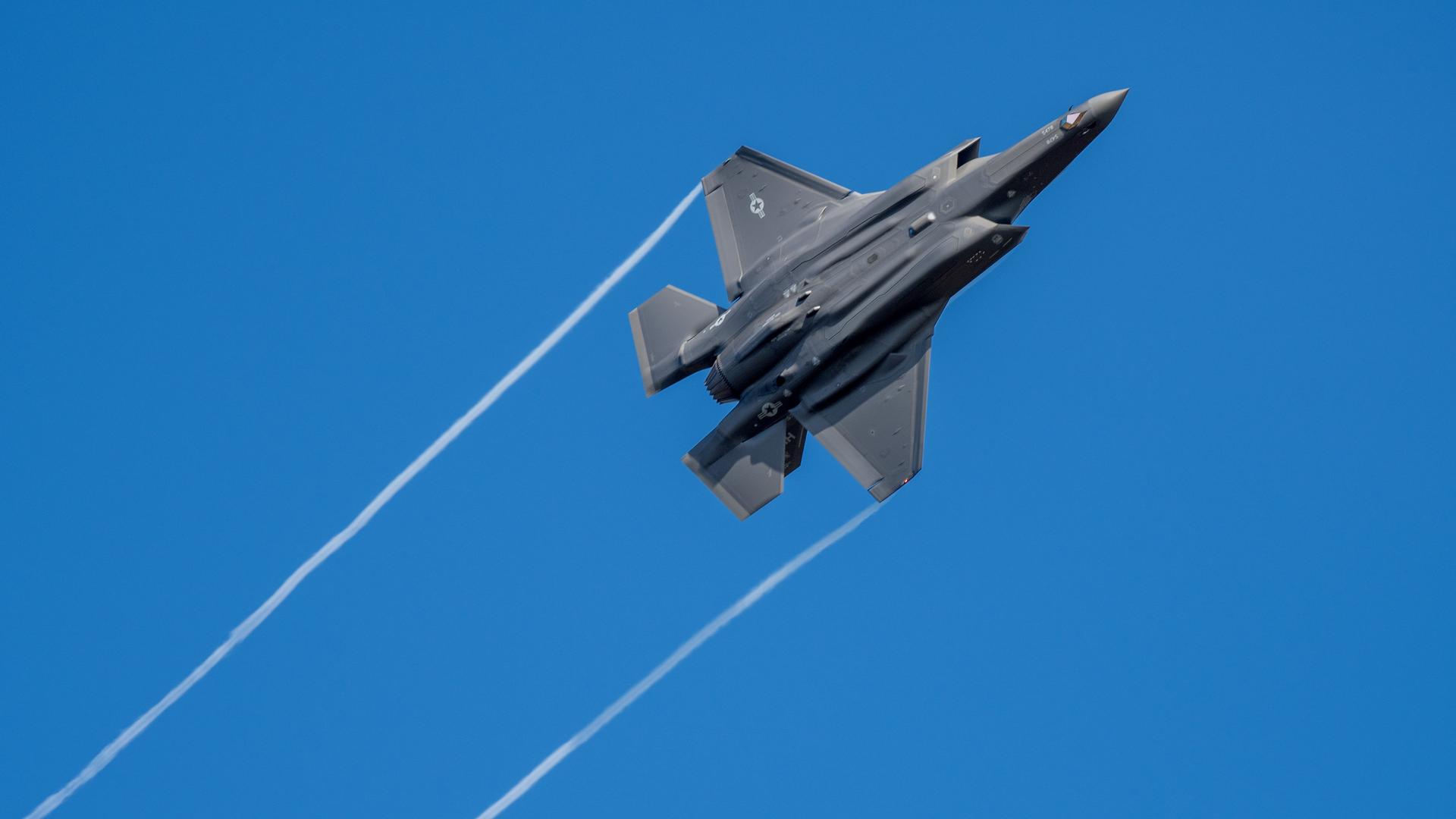 Blick in den Himmel: Man sieht den schwarzen Kampfjet von unten, der durch den blauen Himmel fliegt und von den Tragflächen weiße Kondensstreifen hinterlasst. Blick in den Himmel: Man sieht den schwarzen Kampfjet von unten, der durch den blauen Himmel fliegt und von den Tragflächen weiße Kondensstreifen hinterlasst.