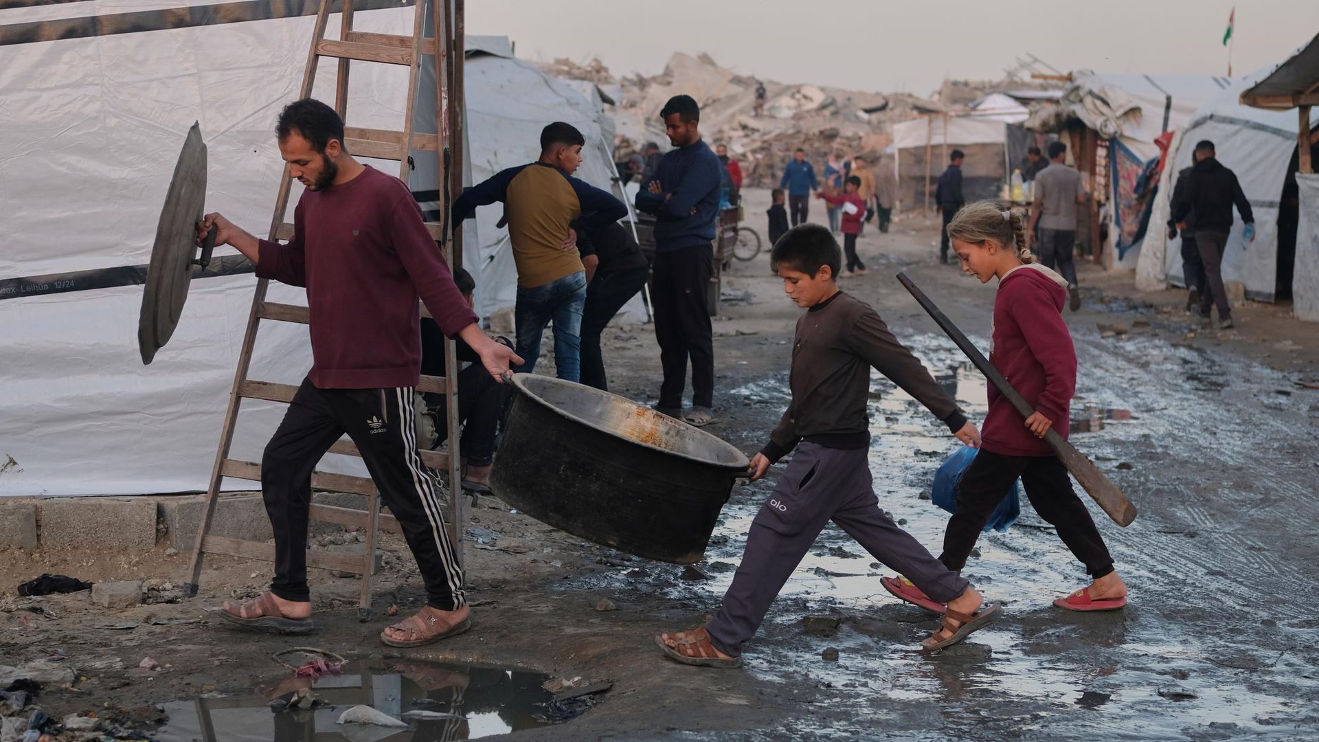 Vertriebene Palästinenser gehen durch schlammige Wege, während ein Mann und Kinder in einem Lager in Jabalia, im nördlichen Gazastreifen, Kochausrüstung tragen. Vertriebene Palästinenser gehen durch schlammige Wege, während ein Mann und Kinder in einem Lager in Jabalia, im nördlichen Gazastreifen, Kochausrüstung tragen.