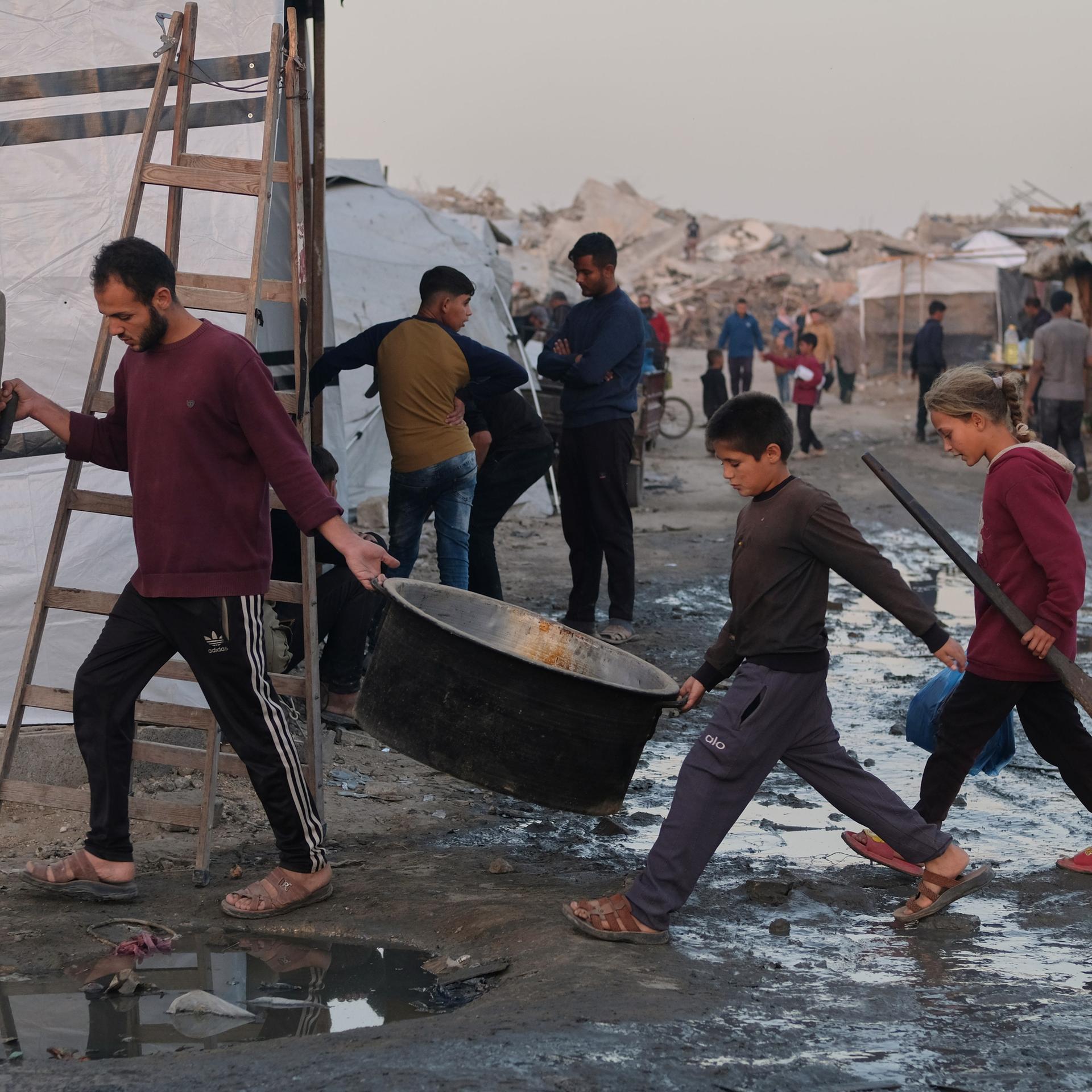 Vertriebene Palästinenser gehen durch schlammige Wege, während ein Mann und Kinder in einem Lager in Jabalia, im nördlichen Gazastreifen, Kochausrüstung tragen.
