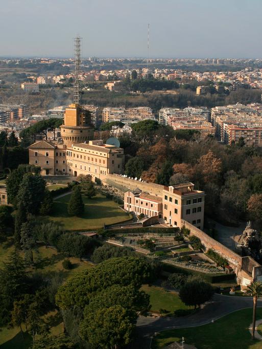 Blick von oben vom Peterstom auf die vatikanischen Gärten. Im Hintergrund steht ein großes Gebäude, dass schmal bergan gebaut ist. Auf einem Turm steht eine hohe Sendeanlage.