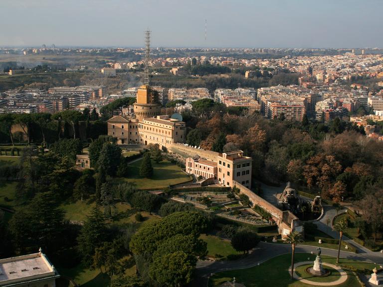 Blick von oben vom Peterstom auf die vatikanischen Gärten. Im Hintergrund steht ein großes Gebäude, dass schmal bergan gebaut ist. Auf einem Turm steht eine hohe Sendeanlage.