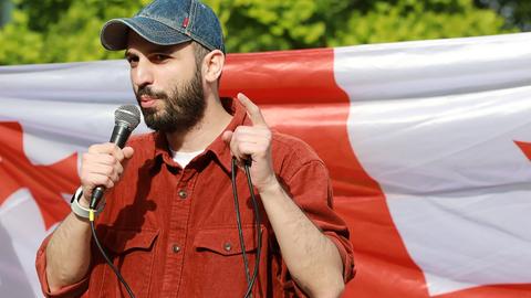 Giorgi Kakabadze steht mit einem Mikrofon in der Hand vor einer georgischen Flagge und spricht