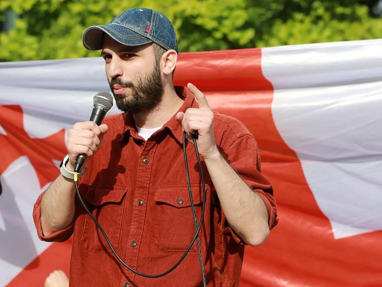 Giorgi Kakabadze steht mit einem Mikrofon in der Hand vor einer georgischen Flagge und spricht