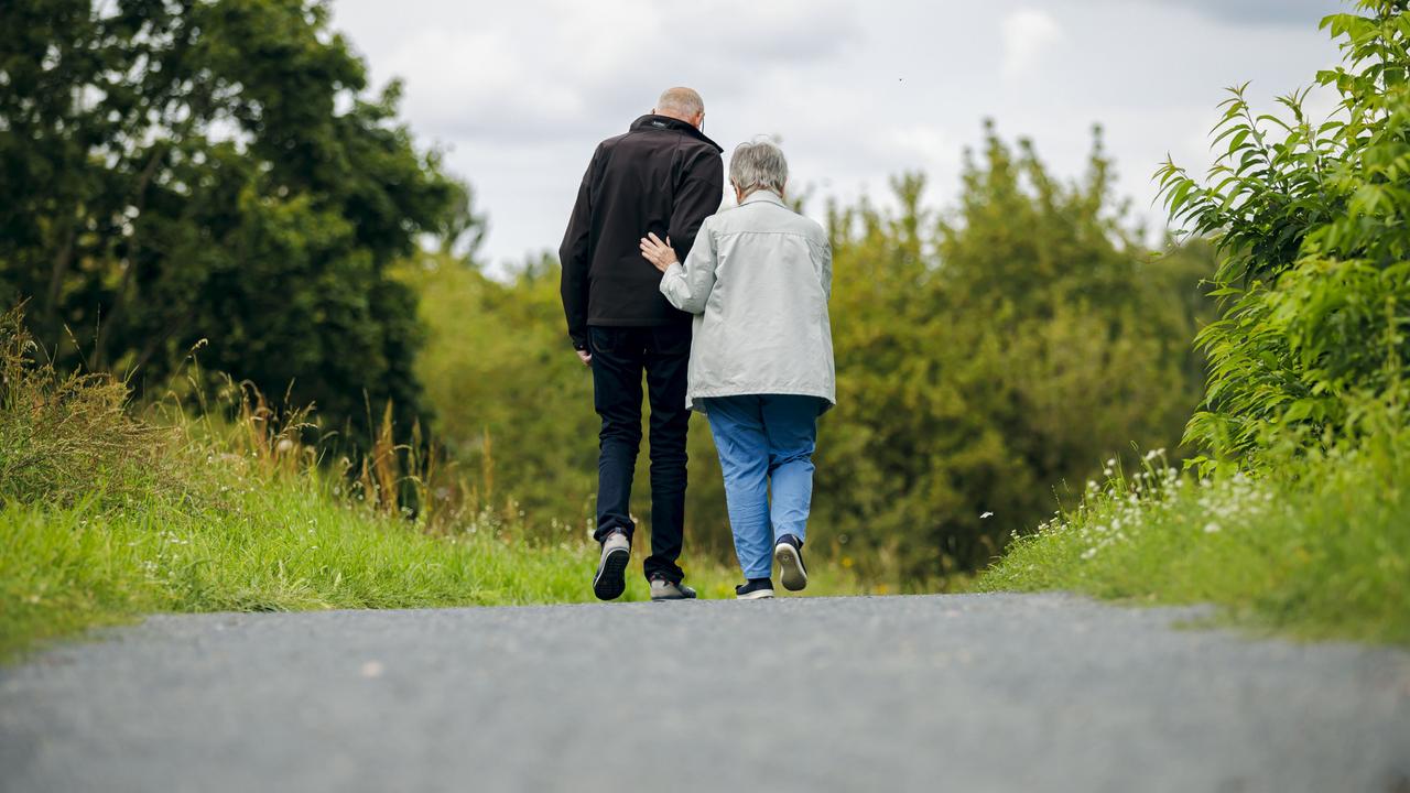 Ein altes Ehepaar läuft bei einem Spaziergang einen Weg entlang, wobei die Frau ihren Mann unterstützt.