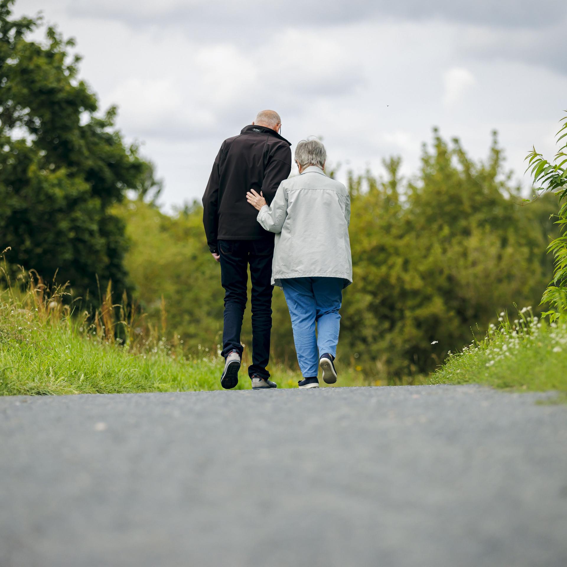 Ein altes Ehepaar läuft bei einem Spaziergang einen Weg entlang, wobei die Frau ihren Mann unterstützt.