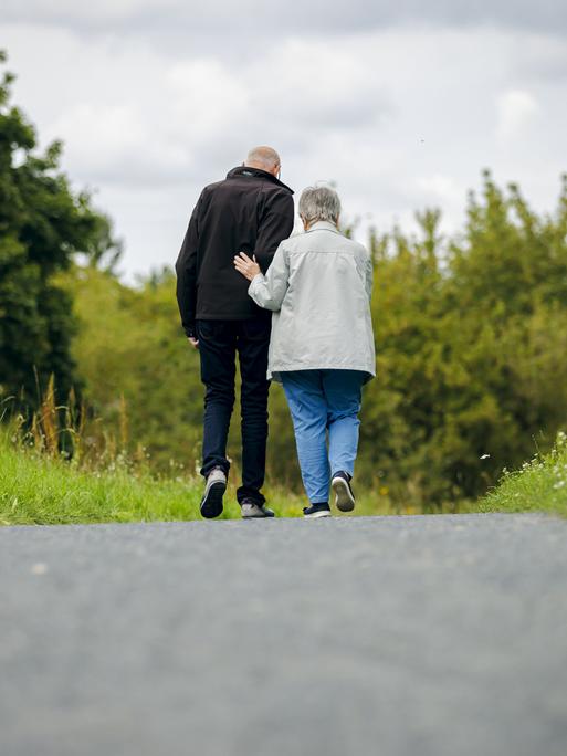 Ein altes Ehepaar läuft bei einem Spaziergang einen Weg entlang, wobei die Frau ihren Mann unterstützt.