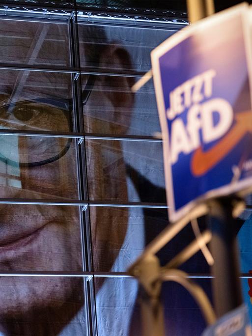 Ein AfD-Plakat hängt an einem Laternenpfahl, im Hintergrund sieht man das Gesicht von Friedrich Merz an einem Gebäude.