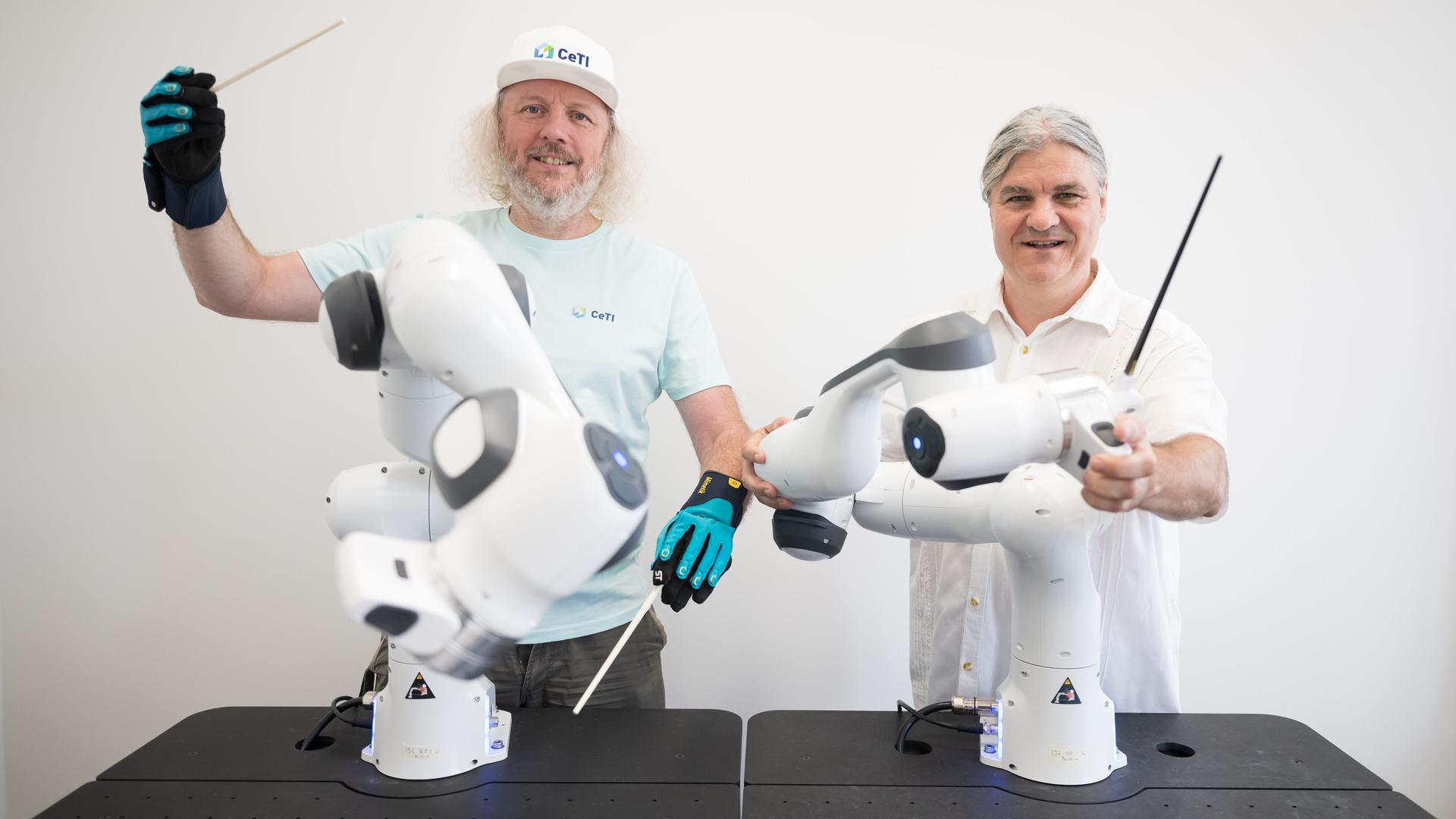 Zwei Männer stehen hinter Roboterarmen. Einer trägt einen Datenhandschuh. Beide Männer haben einen Taktstock in der Hand. 