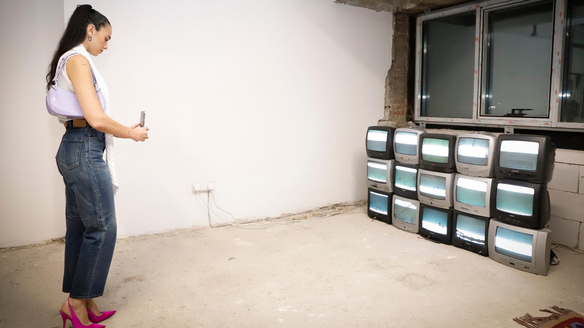 Eine Frau besucht eine Ausstellung. Sie fotografiert mit ihrem Smartphone mehrere Fernsehgeräte die aufeinandergestabelt in einem Raum stehen. Es ist die Popsängerin Dua Lipa, die die Manifesta 14 in Pristina besucht.
