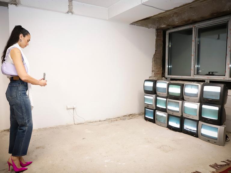 Eine Frau besucht eine Ausstellung. Sie fotografiert mit ihrem Smartphone mehrere Fernsehgeräte die aufeinandergestabelt in einem Raum stehen. Es ist die Popsängerin Dua Lipa, die die Manifesta 14 in Pristina besucht.
