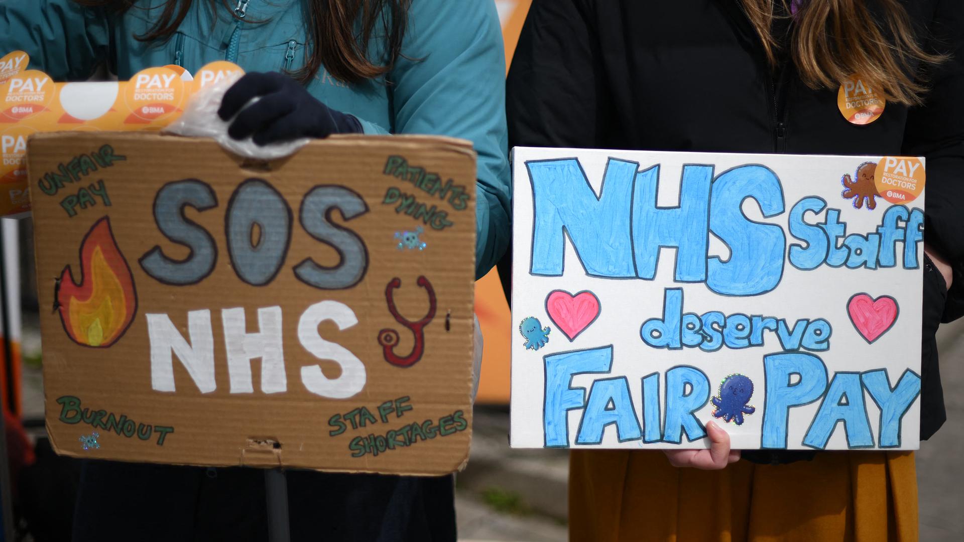 Zwei Mitarbeiterinnen des britischen Gesundheitsdienstes halten Schilder mit der Auschrift "SOS NHS" und "Das NHS-Personal verdient eine faire Bezahlung". Zwei Mitarbeiterinnen des britischen Gesundheitsdienstes halten Schilder mit der Auschrift "SOS NHS" und "Das NHS-Personal verdient eine faire Bezahlung".