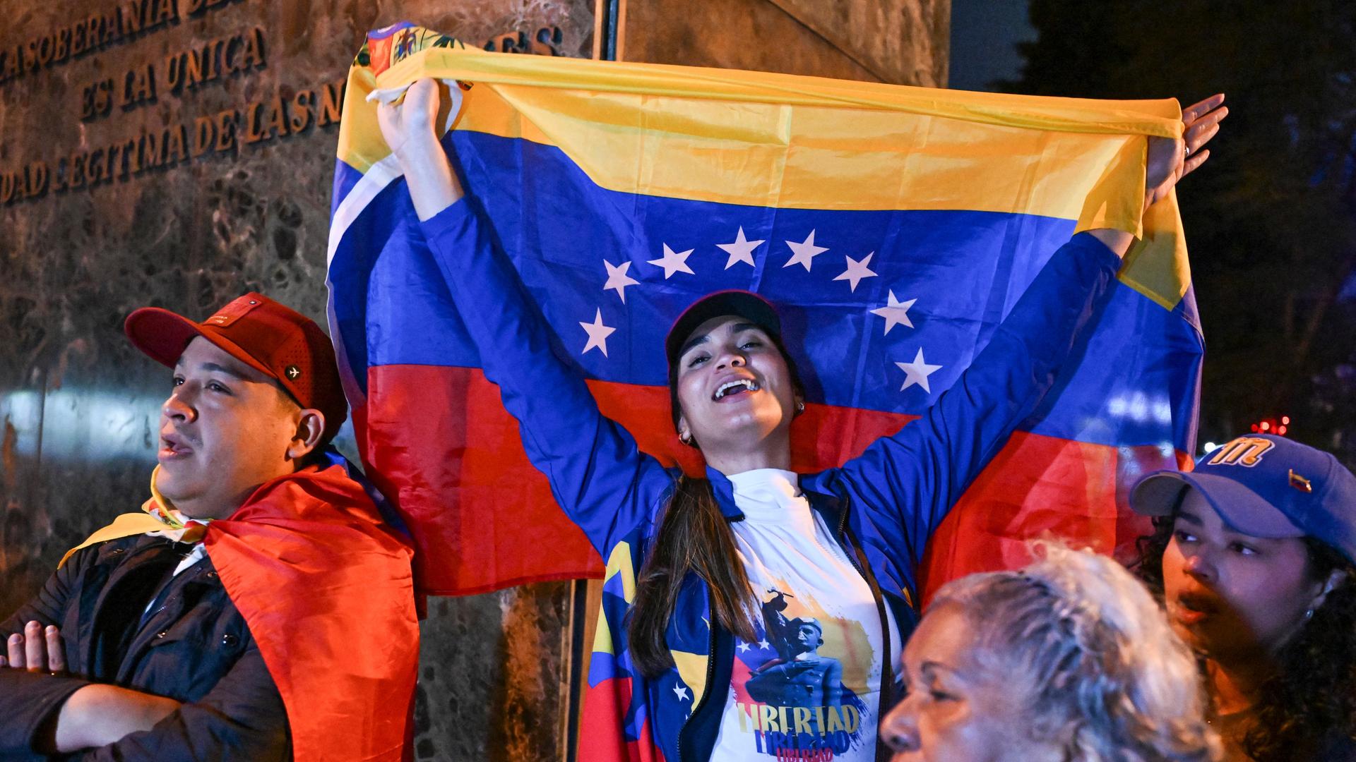 Eine Frau reißt eine venezolanische Nationalflagge in die Höhe. Sie steht zwischen weiteren Teilnehmern einer spontanen Versammlung von Exil-Venezolanern.