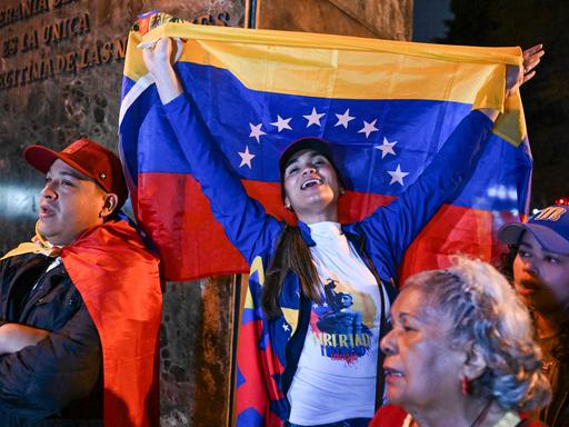 Eine Frau reißt eine venezolanische Nationalflagge in die Höhe. Sie steht zwischen weiteren Teilnehmern einer spontanen Versammlung von Exil-Venezolanern.