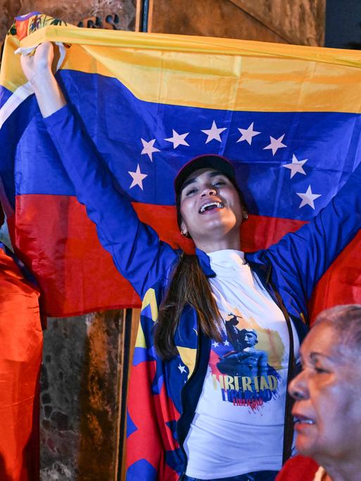 Eine Frau reißt eine venezolanische Nationalflagge in die Höhe. Sie steht zwischen weiteren Teilnehmern einer spontanen Versammlung von Exil-Venezolanern.