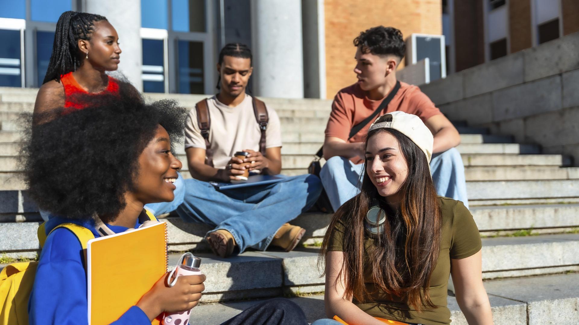 Vielfältige Gruppe junger Studenten, die auf der Treppe vor einem Universitätsgebäude sitzen, sich unterhalten und in der Pause zwischen den Vorlesungen. Vielfältige Gruppe junger Studenten, die auf der Treppe vor einem Universitätsgebäude sitzen, sich unterhalten und in der Pause zwischen den Vorlesungen.