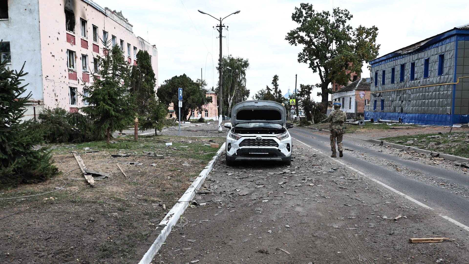 Beschädigte Häuser an einer weitgehend verlassenen Straße. Ein bewaffneter ukrainischer Soldat läuft an einem Auto vorbei.