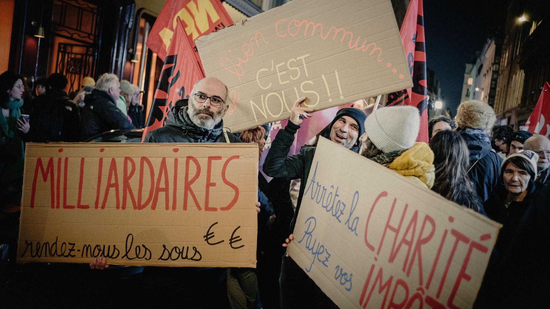 Während einer Protestaktion in Paris halten Menschen Schilder mit den Aufschriften „Milliardäre, gebt uns das Geld zurück, das Gemeinwohl sind wir" und „Hört auf mit der Wohltätigkeit, zahlt eure Steuern".