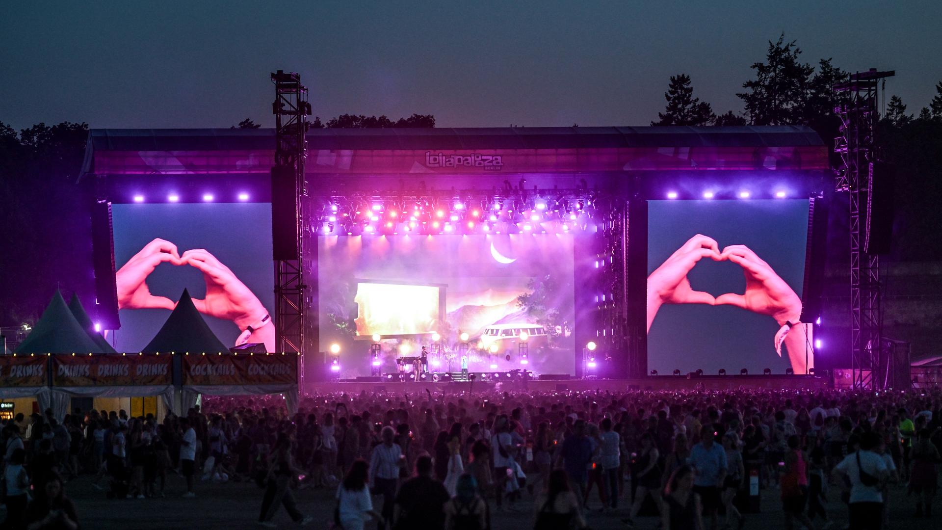 Große Menge auf einem Musikfestival. Auf den Bühnenleinwänden wird gezeigt, wie jemand mit den Händen ein Herz formt.