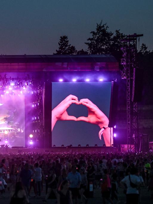 Große Menge auf einem Musikfestival. Auf den Bühnenleinwänden wird gezeigt, wie jemand mit den Händen ein Herz formt.