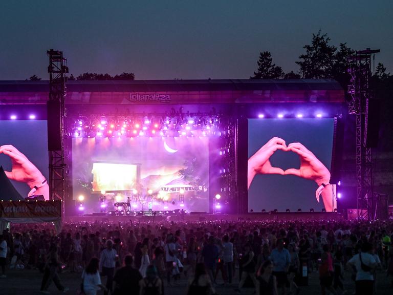 Große Menge auf einem Musikfestival. Auf den Bühnenleinwänden wird gezeigt, wie jemand mit den Händen ein Herz formt.