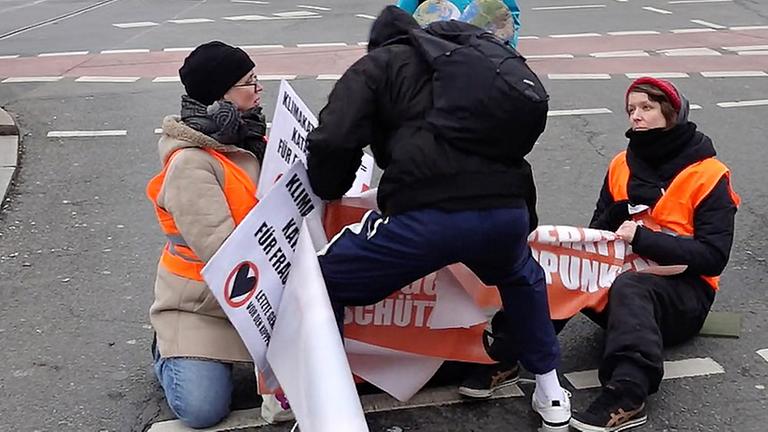 Bei einer Aktion der "Letzten Generation'" blockieren Umweltaktivistinnen eine Kreuzung im Stadtteil Prenzlauer Berg. Ein Mann versucht dabei, ihnen ihre Transparente wegzureißen.
