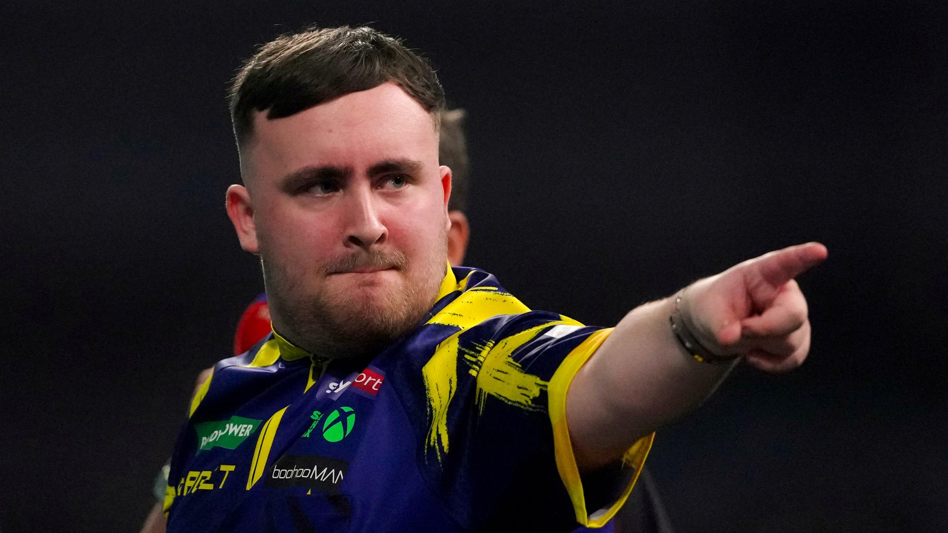 Luke Littler zeigt mit dem Finger nach rechts und schaut jubelnd.