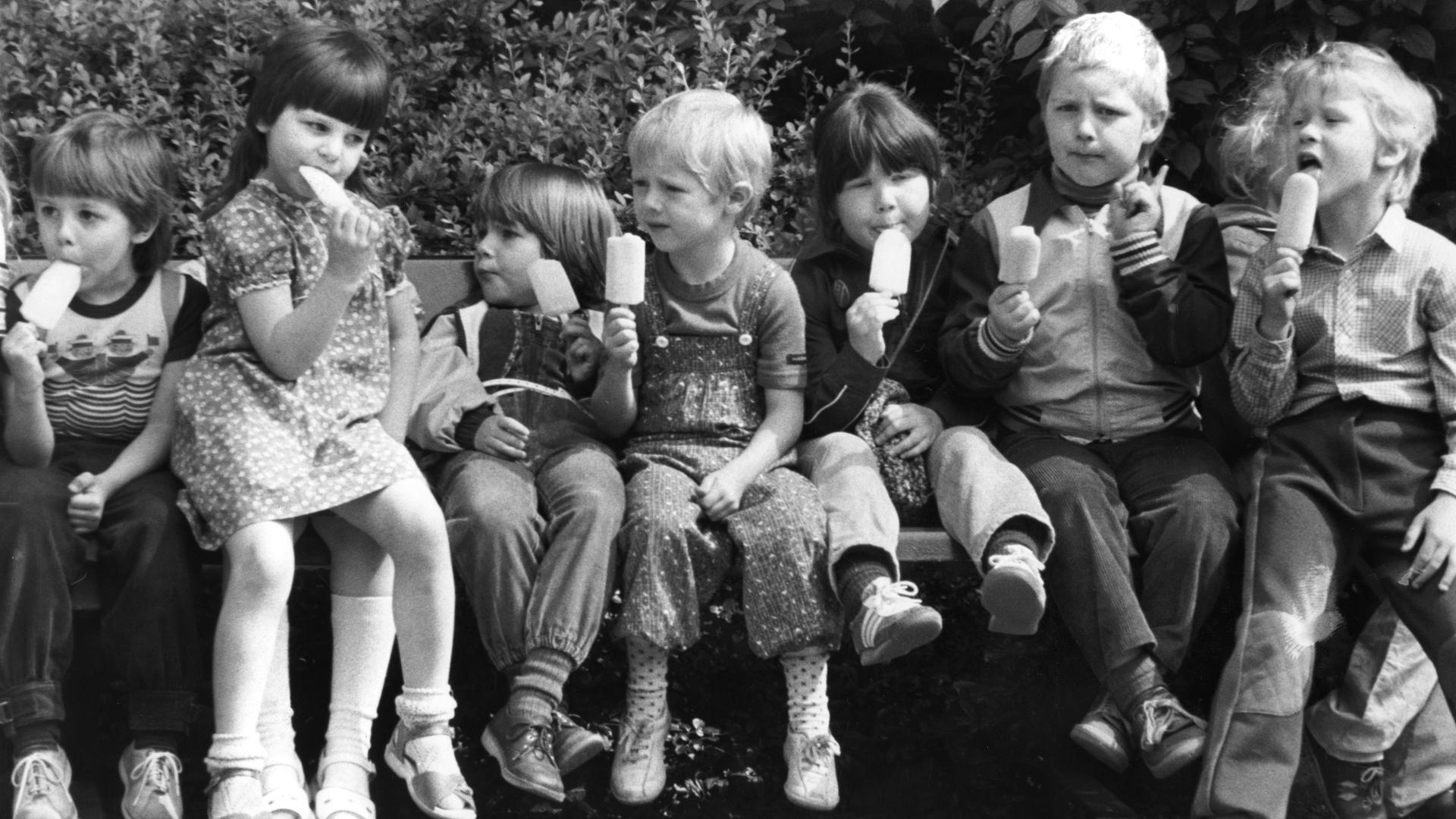 Kinder im Zoologischen Garten in Berlin auf einer Bank und essen Eis am Stiel. (Foto von 1983)