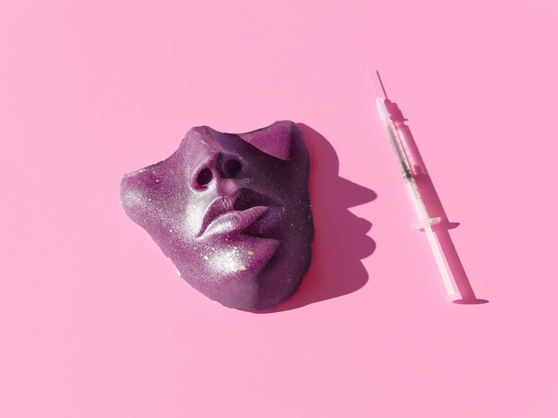 Eine Spritze und eine violette Maske vor pinkem Hintergrund