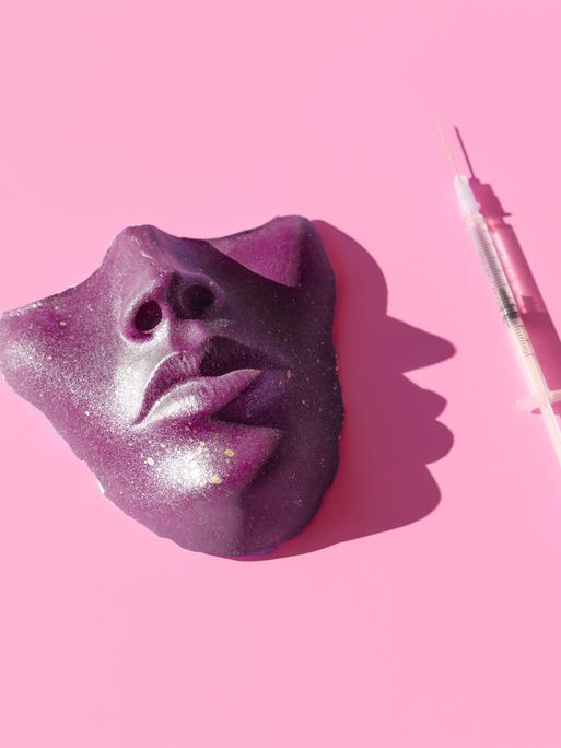 Eine Spritze und eine violette Maske vor pinkem Hintergrund