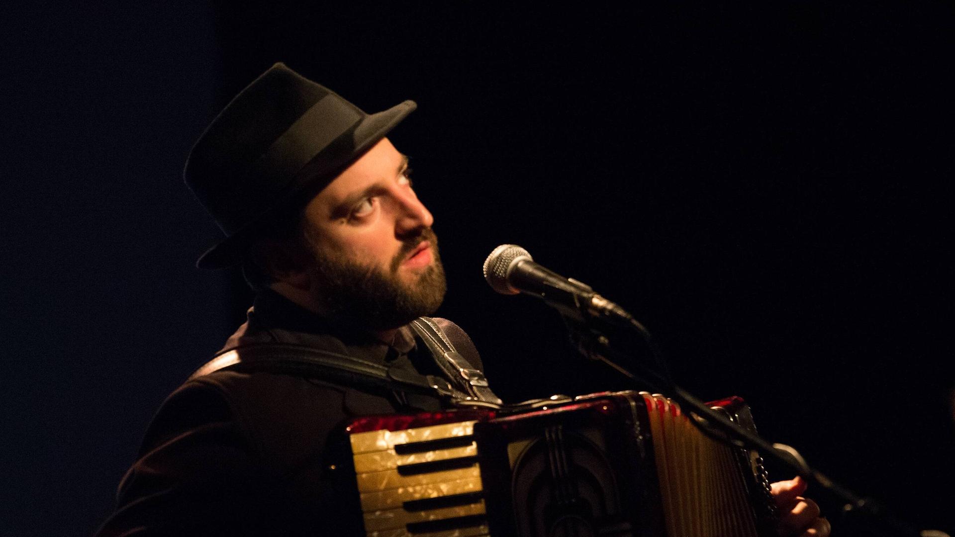 Daniel Kahn mit Hut spielt Akkordeon und singt auf einer Bühne mit Mikrofon.