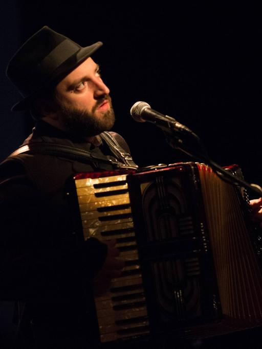 Daniel Kahn mit Hut spielt Akkordeon und singt auf einer Bühne mit Mikrofon.