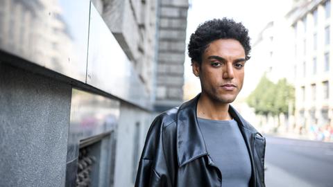 Lamin Leroy Gibba, er trägt eine schwarze Jacke und ein graues T-Shirt. Der Hintergrund ist verschwommen.