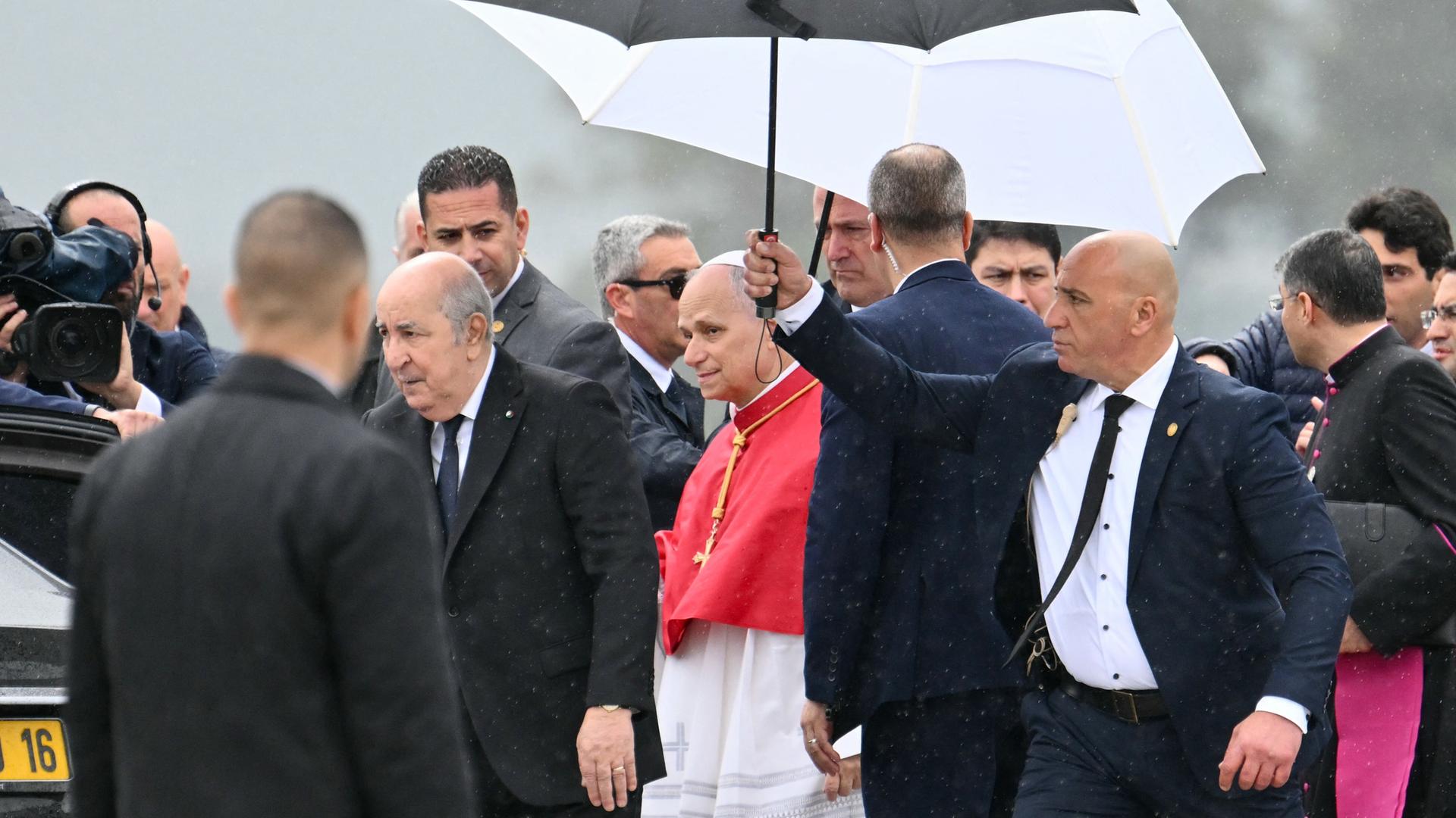 Papst Leo XIV. wird von Algeriens Präsident Tebboune nach seiner Landung auf dem Houari Boumediene International Airport begrüßt. Ein Sicherheitsbeamter hält einen Schirm über ihn.