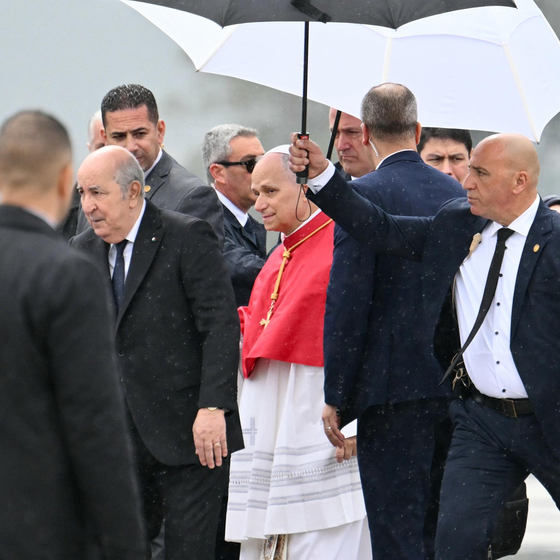 Papst Leo XIV. wird von Algeriens Präsident Tebboune nach seiner Landung auf dem Houari Boumediene International Airport begrüßt. Ein Sicherheitsbeamter hält einen Schirm über ihn.