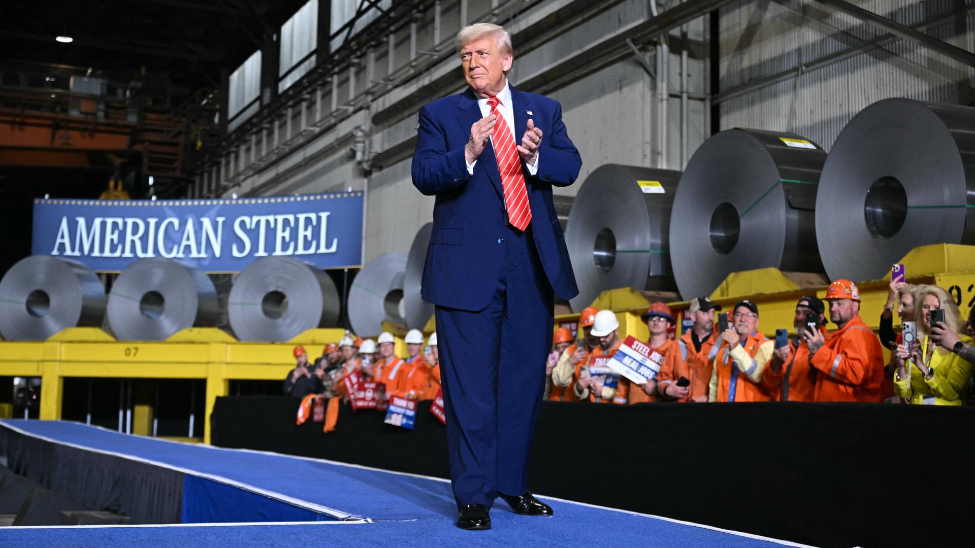 US-Präsident Trump läuft über einen Laufsteg, der mit einem blauen Teppich ausgekleidet ist. An den Seiten stehen Arbeiter, im Hintergrund sieht man große Rollen mit Rohstahl.