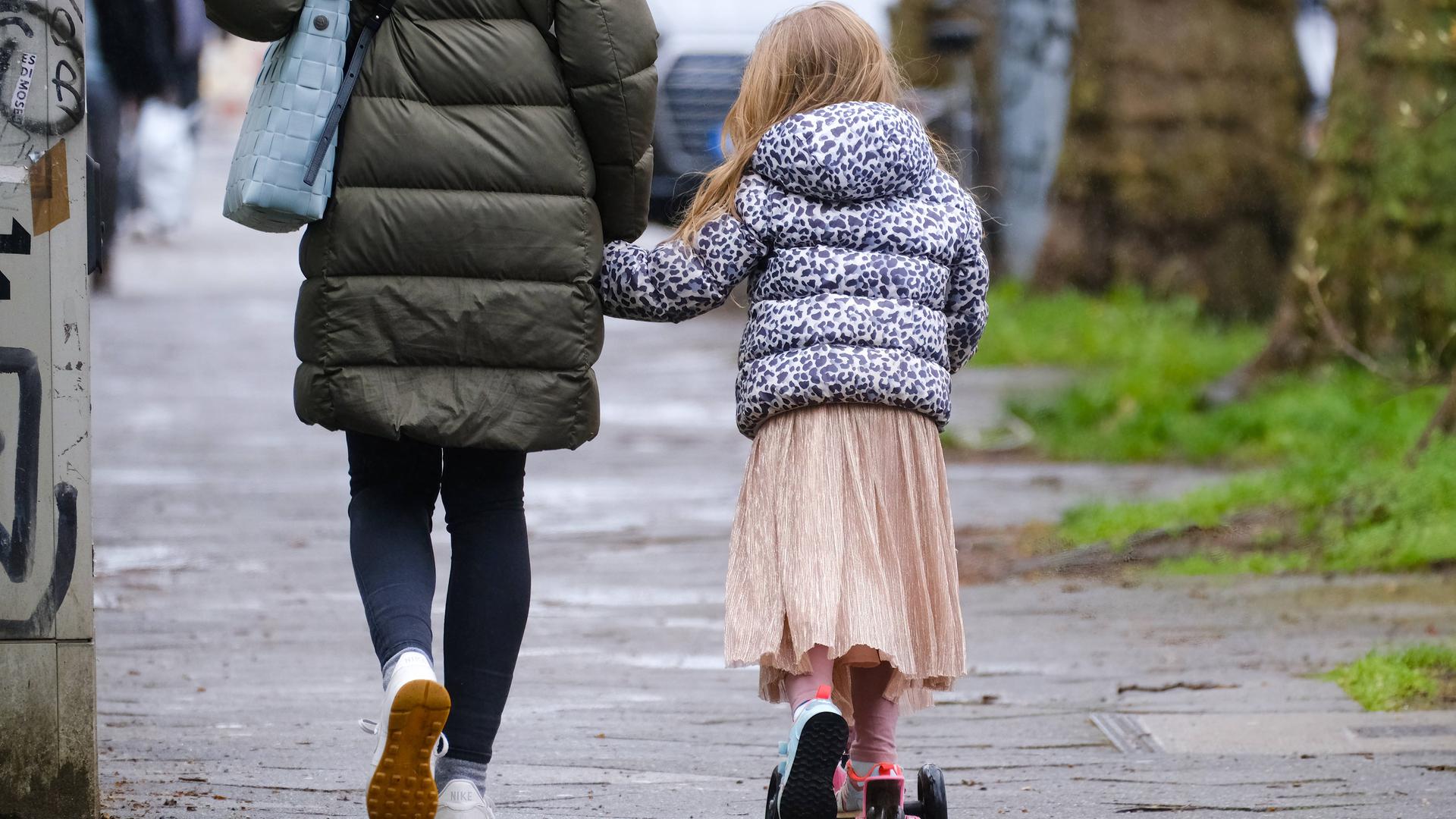 Eine Tochter mit Tretroller läuft neben ihrer Mutter.