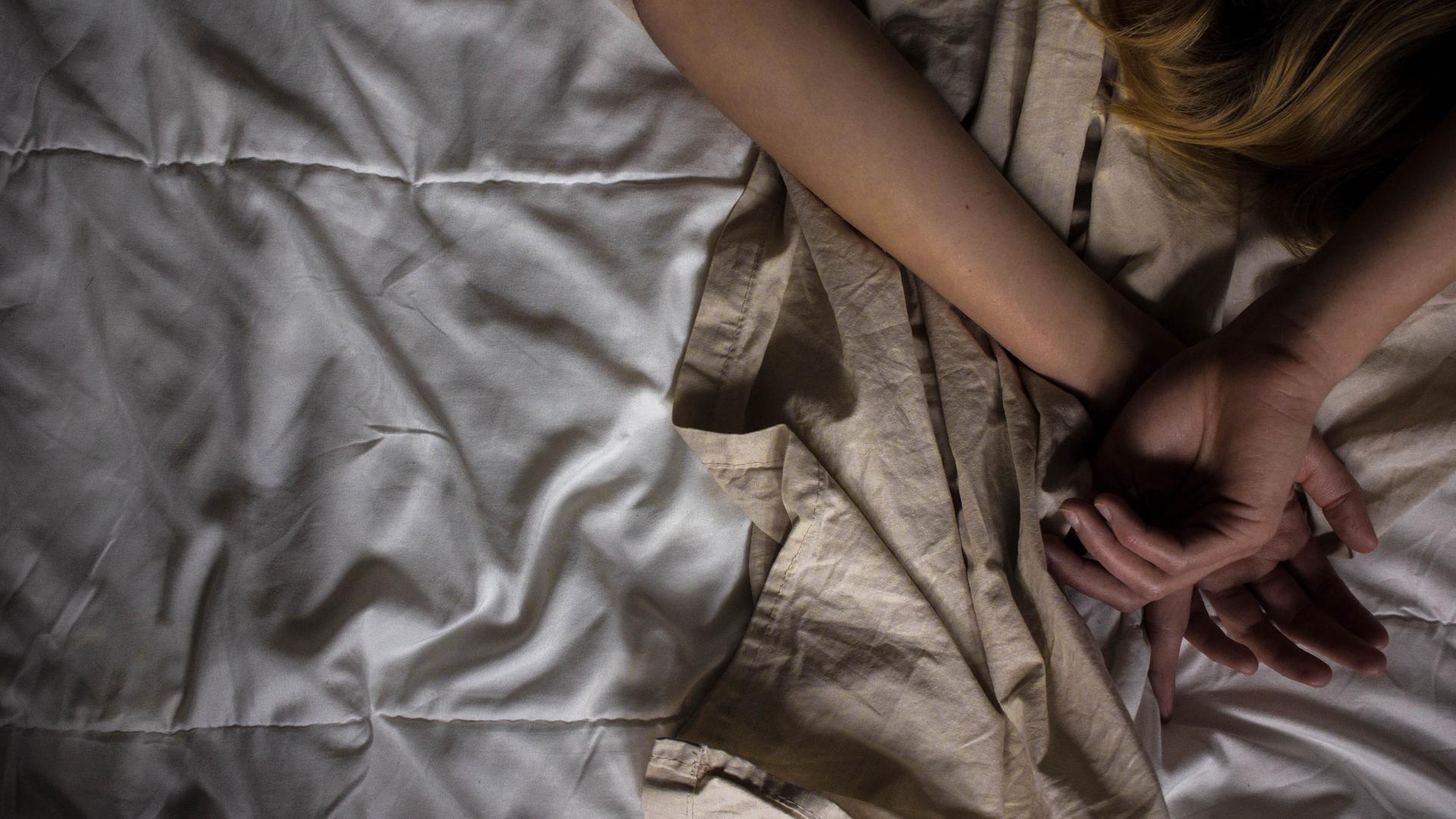 In einem Bett liegt eine Person, die die Arme über ihrem Kopf hat. Im Bild sieht man nur die Arme und einen Teil der Haare. In einem Bett liegt eine Person, die die Arme über ihrem Kopf hat. Im Bild sieht man nur die Arme und einen Teil der Haare.