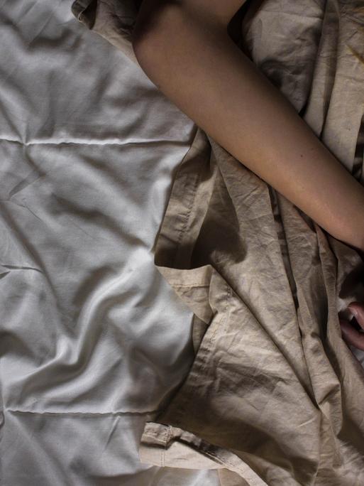 In einem Bett liegt eine Person, die die Arme über ihrem Kopf hat. Im Bild sieht man nur die Arme und einen Teil der Haare.