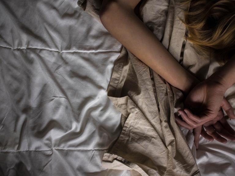In einem Bett liegt eine Person, die die Arme über ihrem Kopf hat. Im Bild sieht man nur die Arme und einen Teil der Haare.