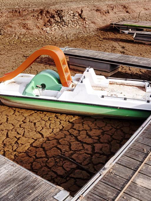 Ein Tretboot mit roter Rutsche liegt an einem Steg auf rissigen Lehm eines ausgetrockneten Sees.