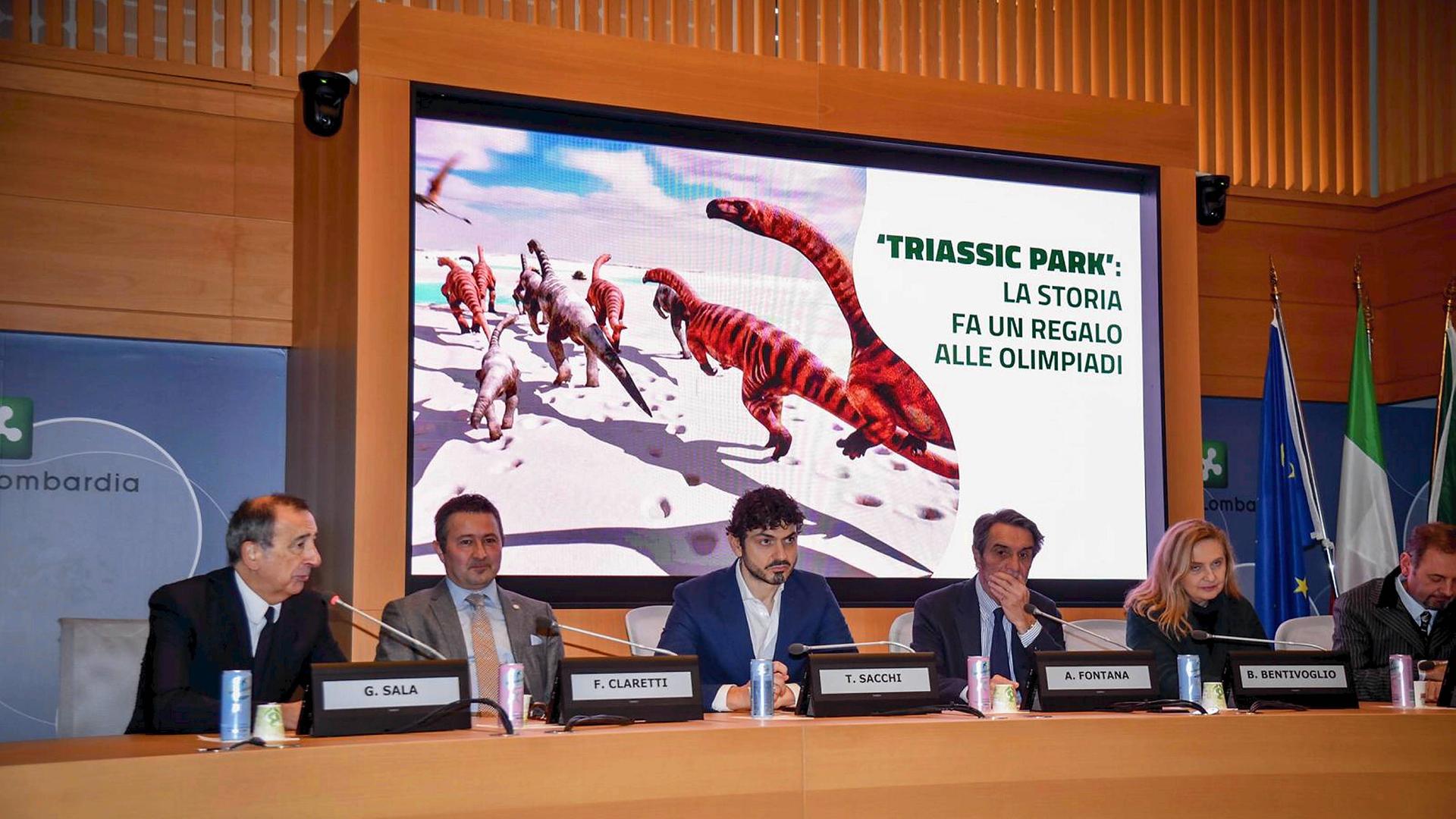 Mehrere Menschen sitzen bei einer Pressekonferenz an einem Tisch mit Mikrofonen. Im Hintergrund ist eine Leinwand mit einer computergenerierten Grafik von Dinosauriern und dem Titel "Triassic Park: Die Geschichte beschenkt die Olympischen Spiele". Mehrere Menschen sitzen bei einer Pressekonferenz an einem Tisch mit Mikrofonen. Im Hintergrund ist eine Leinwand mit einer computergenerierten Grafik von Dinosauriern und dem Titel "Triassic Park: Die Geschichte beschenkt die Olympischen Spiele".